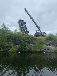 Upside-down car suspended above dark water, crane lifting it.