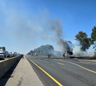 Vehicle fire on highway, black smoke rising, firefighters present, traffic backed up