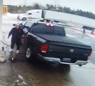 Black pickup truck at a gas station. A person in a dark jacket and pants is fueling the vehicle. A semi-truck is visible in the background.