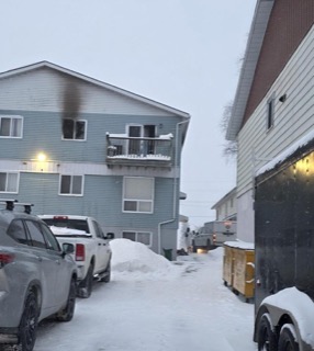 Snow-covered street with parked cars and a blue apartment building.
