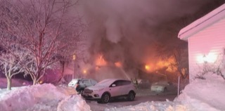 Snow-covered street with parked cars and a house with visible smoke.