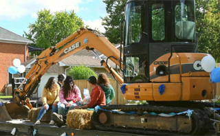 Four people sitting on hay bales in front of an excavator.