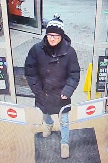 Person wearing a dark coat, knit hat, and glasses, standing in a doorway.