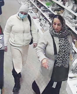 Two people walking in a store, one wearing a light-colored puffer jacket and a face mask, the other wearing a leopard-print scarf and a black shirt