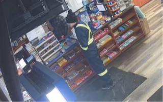 Person in dark clothing and a yellow safety vest standing in a store, shelves stocked with products visible