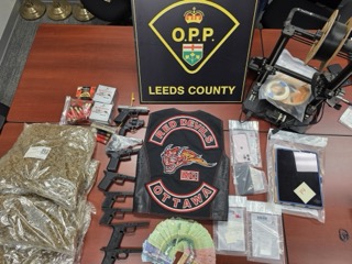 Firearms, ammunition, cash, and a patch reading &quot;Red Devils Ottawa MC&quot; displayed on a table with the Ontario Provincial Police and Leeds County logos.