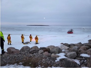 Rescue workers in yellow suits assisting someone on a snow-covered shoreline.