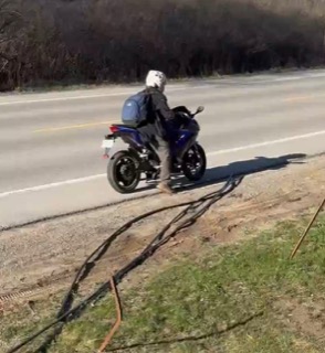 Motorcyclist wearing a helmet and backpack, facing away from the camera, on a paved road.
