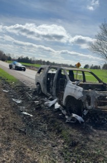 Burned car on the side of a road, police vehicle in the background.