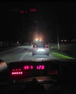 Dashboard view, speedometer reads 120, police car ahead with flashing lights