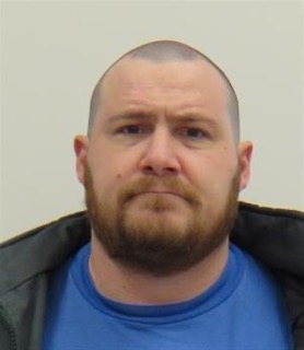 Man with a shaved head and beard, wearing a black jacket and blue shirt.