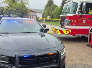 Police cruiser with emergency lights activated, fire truck in background, yellow caution tape visible