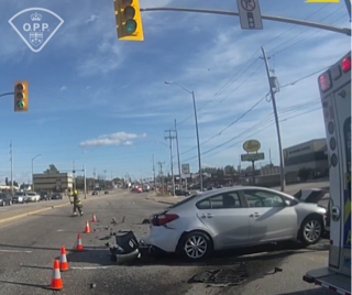 Damaged silver sedan, debris scattered on road, emergency vehicle in background, traffic signal overhead
